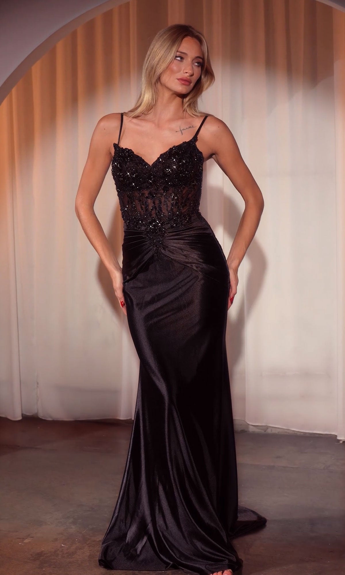 A woman stands indoors in the Beaded-Bodice Long Prom Dress (CLCD776), a sleek black gown with thin straps, a semi-sheer beaded bodice, and a floor-length silhouette.